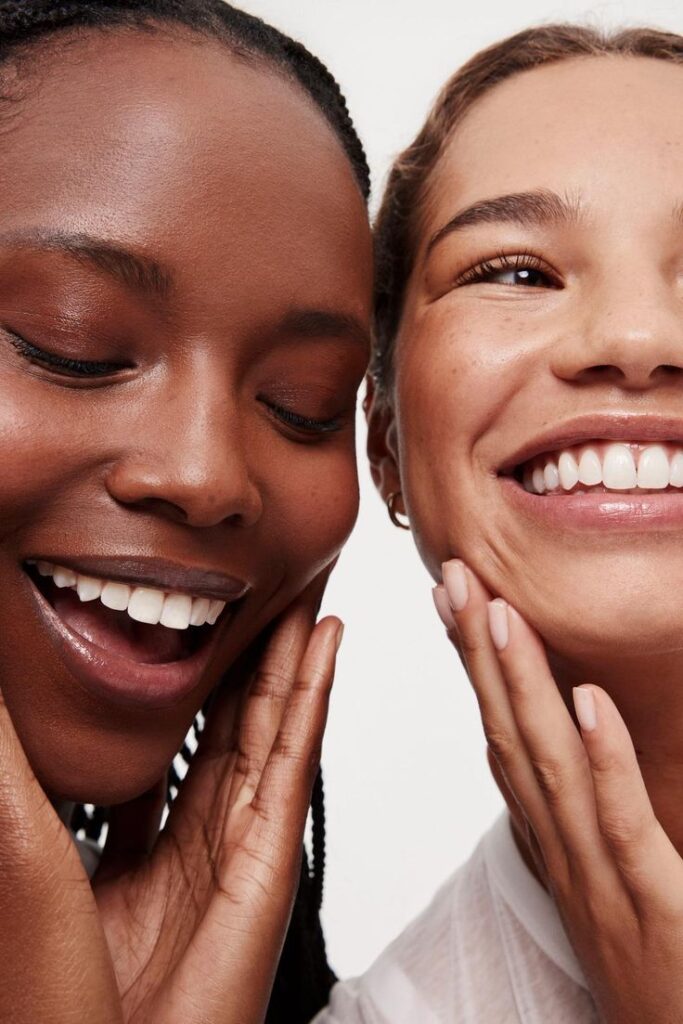 imagem com duas mulheres que representam os tipos de tom de pele e subtom de pele. uma mulher é branca e a outra é negra. a foto mostra parte do rosto delas, elas estão sorrindo.