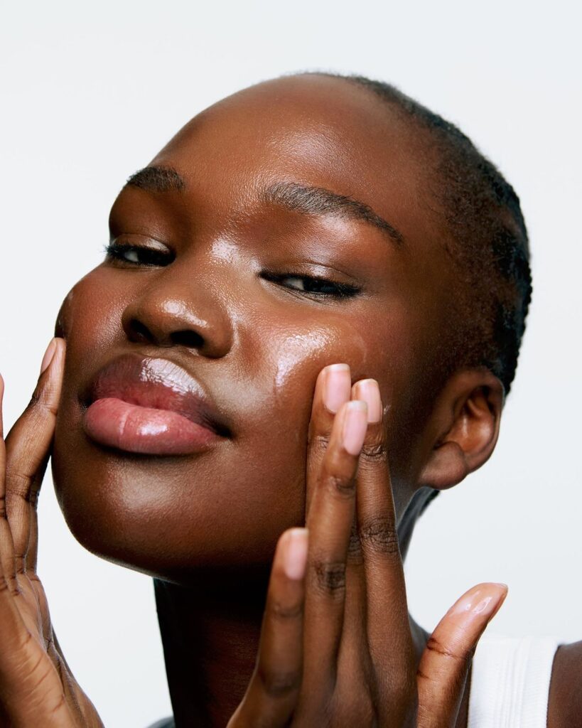 Rosto de mulher negra, com a expressão relaxada, mãos no rosto, e espalhando um hidratante facial.