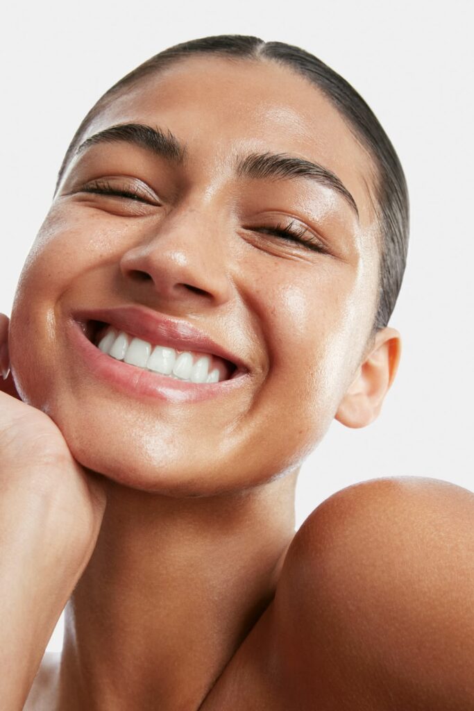 Rosto de mulher morena, sorrindo, com uma das mãos no rosto, e usando hidratante facial.