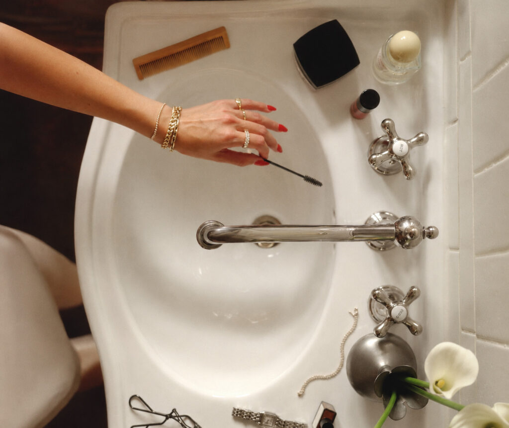Imagem mostra uma pia de cima, produtos de maquiagem e skincare no entorno e uma mão de uma mulher com unhas vermelhas segurando uma escovinha de sobrancelhas. Ela representa a importância de limpar esponja de maquiagem.