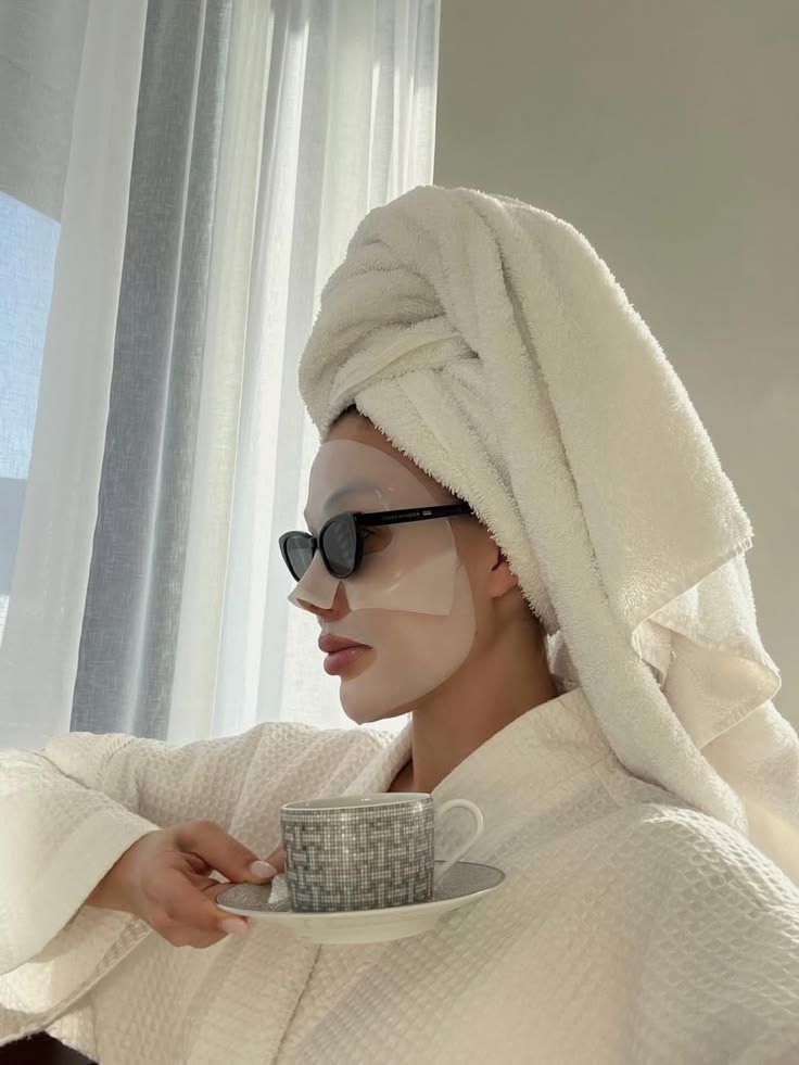 Mulher com máscara facial, óculos escuros, toalha na cabeça, roupão e xícara na mão representando os cuidados com a pele no frio.